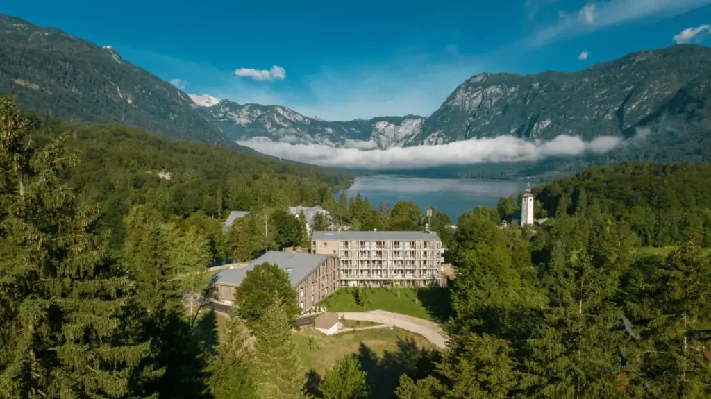 A high-angle view of the modern timber-framed Hotel Bohinj nestled in the Julian Alps, featuring sustainable larch architecture by OFIS Architects overlooking Lake Bohinj and the Church of St. John the Baptist.