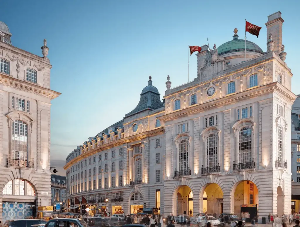 The historic white Portland stone facade of Hotel Café Royal London on Regent Street, a Grade II listed landmark featuring iconic arched entryways and the building's distinctive copper-domed roof against a twilight sky.