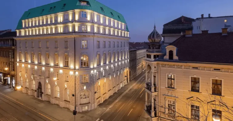 The illuminated early 20th-century Art Deco facade of Hotel Capital Zagreb at night, showing the historic mint-green mansard roof and classical stone arches on a central street corner.