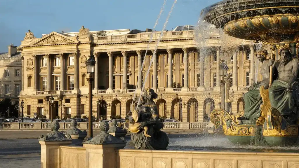 The neoclassical facade of Hôtel de Crillon in Paris, an 18th-century palace commissioned by King Louis XV, viewed from Place de la Concorde behind the ornate Fontaines des Mers at golden hour.