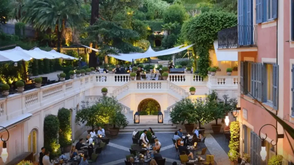 An evening view of the Hotel de Russie Rome "Secret Garden" in Rome, showing guests dining on the tiered Mediterranean terrace surrounded by lush greenery and illuminated neoclassical stone balustrades.