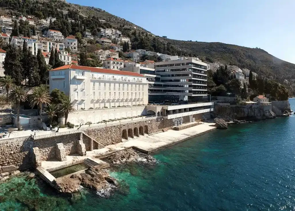 Aerial view of Hotel Excelsior Dubrovnik showing the historic white stone Villa Odak and the modern glass tower integrated into a cliffside with a private beach and rock-cut swimming pool on the Adriatic Sea.