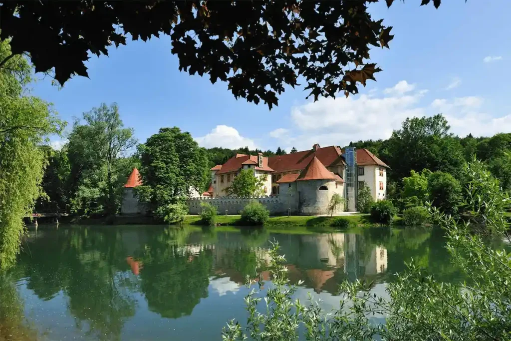 A view of Hotel Grad Otočec, a medieval Gothic castle situated on a small island in the middle of the Krka River in Slovenia, featuring stone walls, red-tiled circular towers, and lush green trees reflecting in the calm emerald water.
