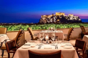 A luxury dinner setting at the rooftop of the Hotel Grande Bretagne Athens, a historic 19th-century palace that hosted King Edward VII's state banquets, showing white-clothed tables with a direct, illuminated view of the Acropolis and Parthenon.