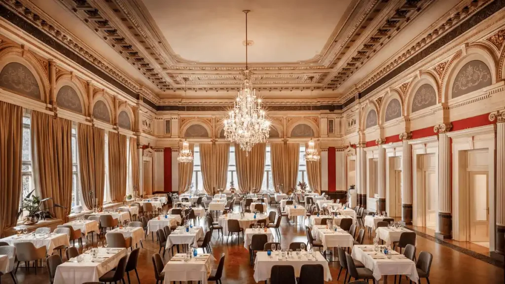 The interior of the historic Golden Hall at Hotel Imperial Opatija, featuring high ornate ceilings, grand crystal chandeliers, and classical stucco details arranged for fine dining.