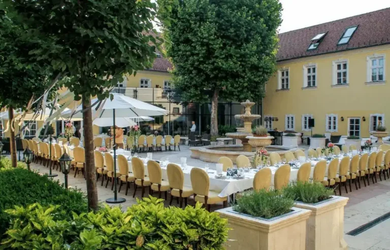 The serene late-baroque inner courtyard of Hotel Kristály Imperial in Tata, featuring an elegant outdoor banquet setup with white linens and a classic stone fountain.