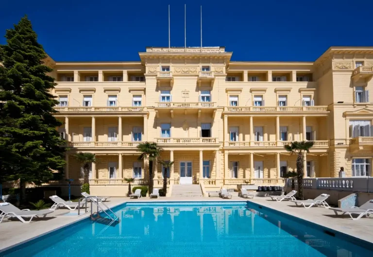 The grand neoclassical facade of Hotel Kvarner Opatija featuring an outdoor swimming pool, elegant columns, and sun loungers under a clear blue sky.