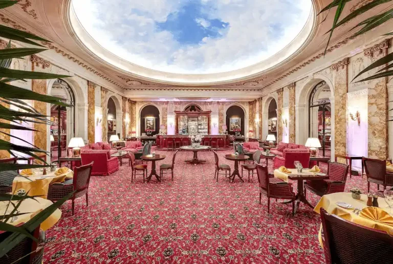 The grand L'Esterel lounge at Hotel Le Plaza Brussels featuring a circular trompe-l'œil sky ceiling, marble columns, and 1930s-inspired red velvet seating.