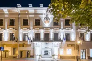 The illuminated 17th-century facade of Hotel Pacai Vilnius at night, featuring the noble Pacas family coat of arms and the arched entrance to the private Baroque courtyard.
