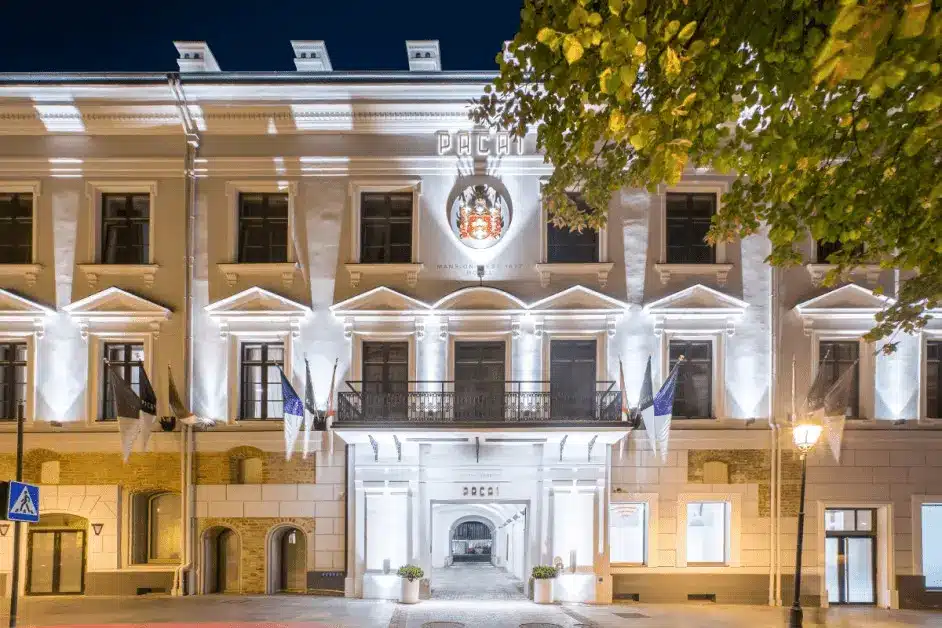 The illuminated 17th-century facade of Hotel Pacai Vilnius at night, featuring the noble Pacas family coat of arms and the arched entrance to the private Baroque courtyard.