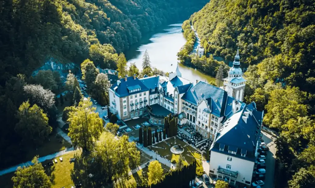 An aerial view of Hotel Palota Lillafüred, a grand Neo-Renaissance castle hotel in Hungary, featuring its iconic pointed turrets and stone facades nestled within the dense forests of the Bükk Mountains alongside the tranquil Lake Hámori.
