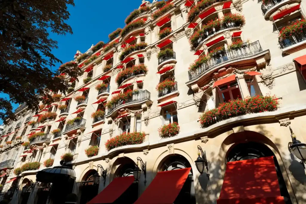 The iconic Haussmann-style facade of Hôtel Plaza Athénée Paris, featuring its signature bright red awnings and flower-filled window boxes against the limestone exterior on Avenue Montaigne.