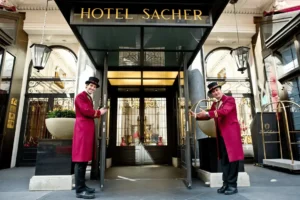 Two doormen in traditional red coats standing at the grand entrance of Hotel Sacher Wien, a 19th-century Neo-Renaissance palace located directly opposite the Vienna State Opera.