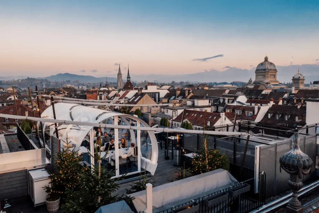 Guests dining inside a transparent Tubbo dome on the Sky Terrace at Hotel Schweizerhof Bern, featuring panoramic winter views of the Bern Old Town and the Federal Palace.