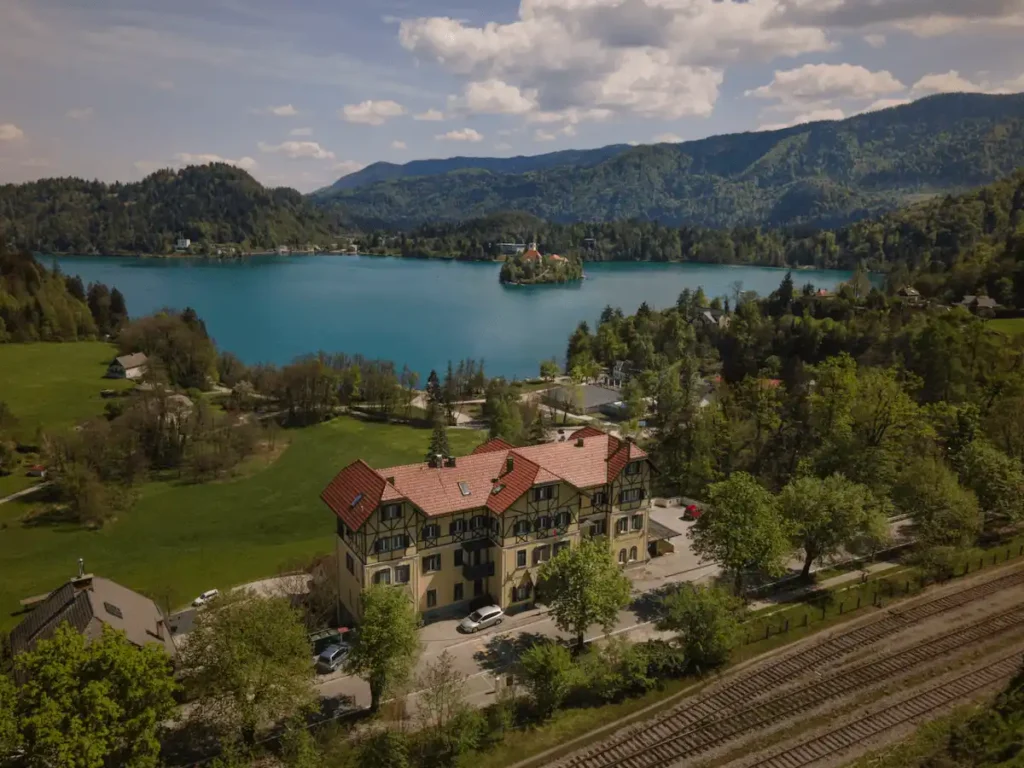 An aerial view of the historic Hotel Triglav Bled, showing its traditional Alpine Secessionist architecture and red-tiled roof, situated on a hill overlooking Lake Bled and the iconic Bled Island.