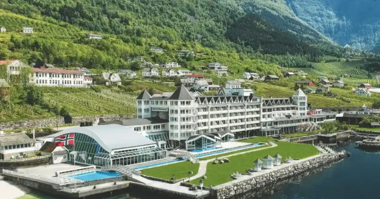 A panoramic view of Hotel Ullensvang situated on the edge of the Hardangerfjord in Lofthus, Norway, featuring its sprawling wings and outdoor infinity pool against a backdrop of snow-capped mountains and lush fruit orchards.