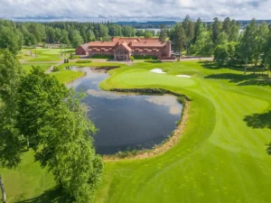 An aerial view of Hotel Vanajanlinna, a grand early 20th-century red-brick manor estate in Hämeenlinna, Finland, featuring its sprawling architectural wings and the lush green fairways of the Linna Golf course surrounding a scenic pond.