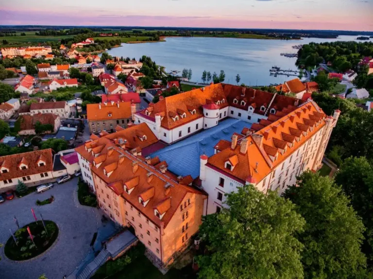 An aerial sunset view of Hotel Zamek Ryn, a massive 14th-century Teutonic Knights' castle in Poland featuring four wings, a large inner courtyard, and orange-tiled roofs, situated between Lake Ołów and Lake Ryńskie in the Masurian Lake District.