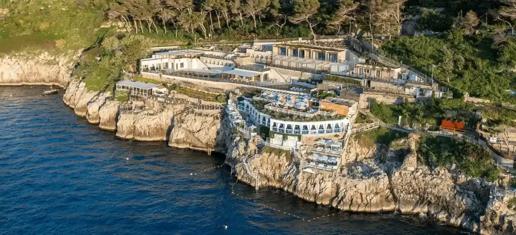 An expansive aerial view of the white, terraced architecture of Jumeirah Capri Palace perched on the rugged limestone cliffs of Anacapri, overlooking the deep blue Mediterranean Sea.