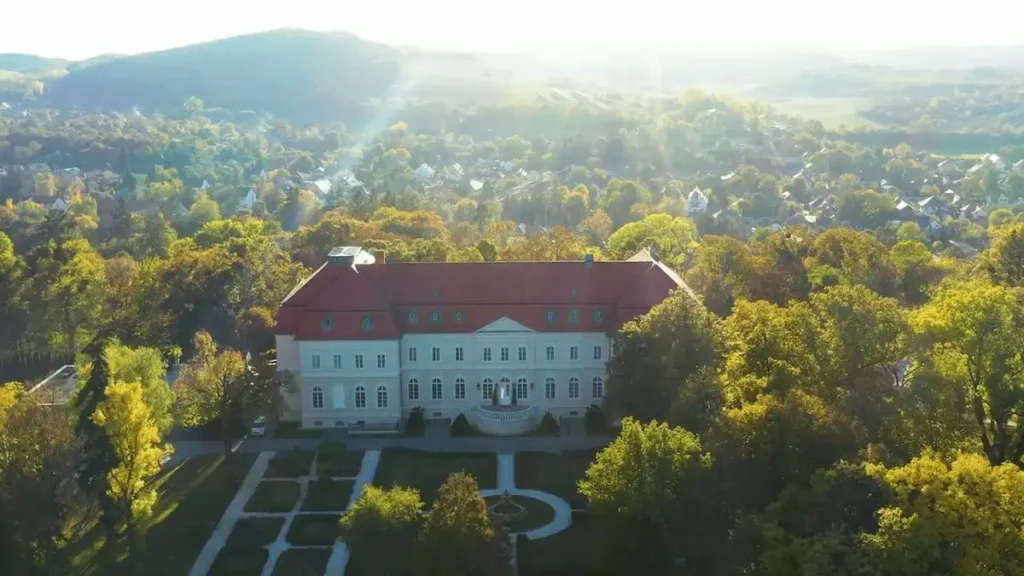 An aerial view of La Contessa Castle Hotel, a grand 19th-century Neo-Classicist mansion in Szilvásvárad, featuring a long symmetrical facade with a red roof and formal manicured gardens, surrounded by the lush green forests of the Bükk Mountains.