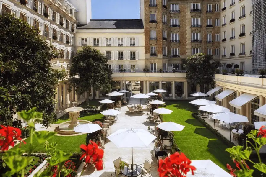 An overhead view of the lush, manicured garden courtyard at Le Bristol Paris, featuring white café umbrellas, a stone fountain, and the hotel's classic French architecture.