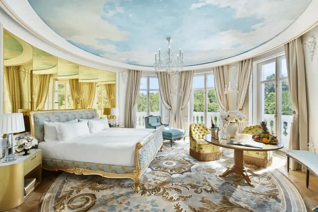 The luxurious Royal Suite at Mandarin Oriental Ritz Madrid, featuring a stunning circular sky-blue ceiling fresco, gilded neoclassical furniture, and floor-to-ceiling windows overlooking the Prado Museum district.