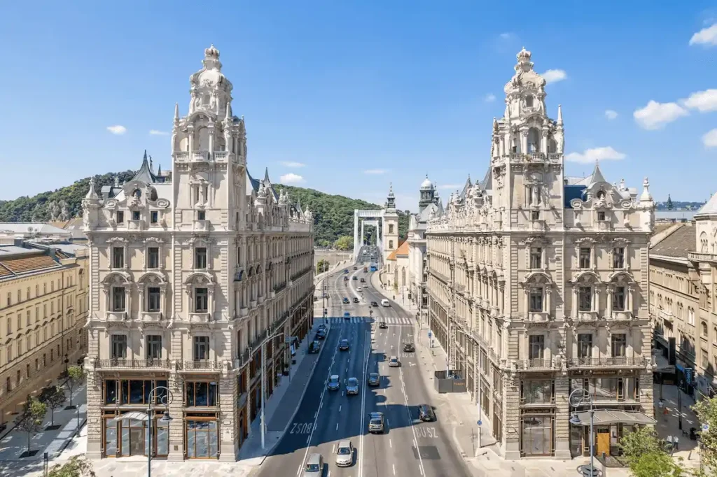 A wide aerial view of the neo-baroque twin buildings of Matild Palace Budapest and Klotild Palace, flanking the entrance to the Elisabeth Bridge under a clear blue sky.