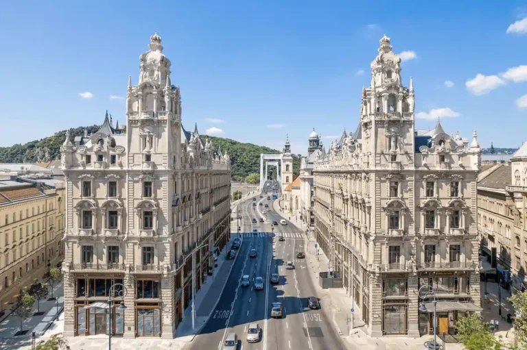 A wide aerial view of the neo-baroque twin buildings of Matild Palace Budapest and Klotild Palace, flanking the entrance to the Elisabeth Bridge under a clear blue sky.