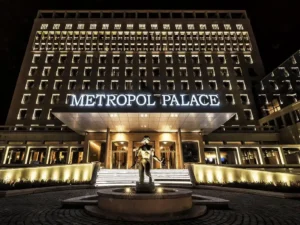 The illuminated modernist facade of Metropol Palace Belgrade at night, featuring the grand entrance, lit-up windows, and the iconic bronze fountain sculpture in the foreground.