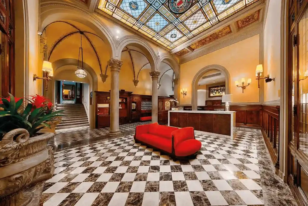 The magnificent lobby of NH Collection Firenze Porta Rossa, featuring a checkerboard marble floor, original Renaissance arches, and a spectacular 19th-century stained-glass ceiling vault.