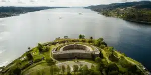 An aerial view of Oscarsborg Castle Hotel, showing the historic circular stone fortress of Oscarsborg situated on an island in the middle of the Oslofjord, surrounded by green coastal vegetation and defensive batteries.