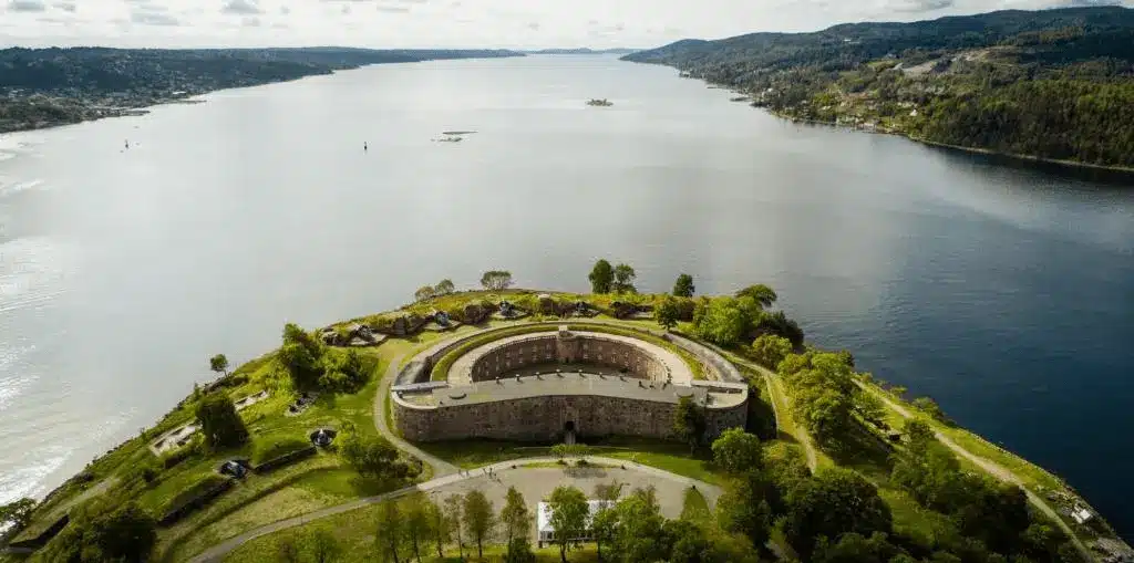 An aerial view of Oscarsborg Castle Hotel, showing the historic circular stone fortress of Oscarsborg situated on an island in the middle of the Oslofjord, surrounded by green coastal vegetation and defensive batteries.