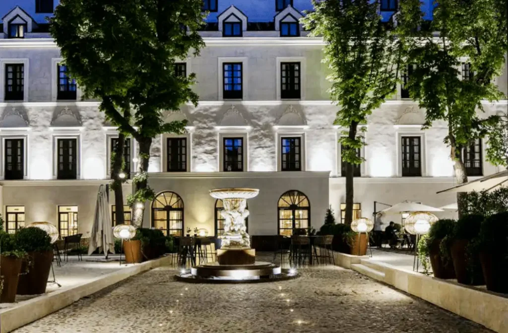 The illuminated 19th-century facade and historic "secret garden" of Palacio de los Duques Gran Meliá, featuring a central stone fountain and lush greenery on the site of a former 13th-century Dominican convent.