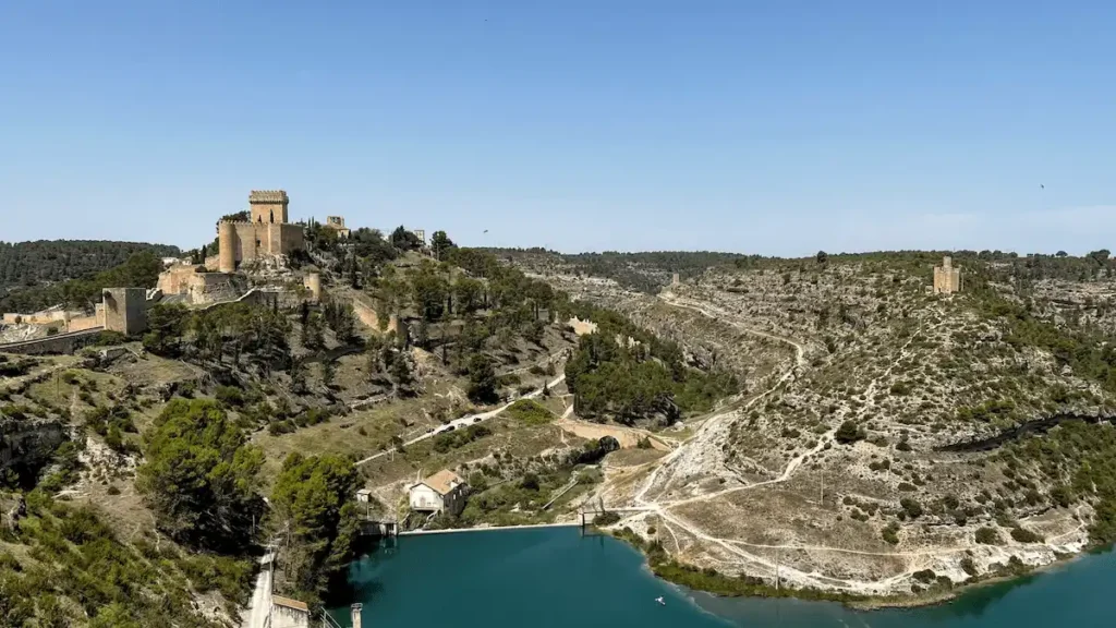 A wide landscape view of the Parador de Alarcón, an 8th-century medieval fortress with a massive square stone keep, situated on a craggy hilltop surrounded by defensive walls and the deep blue waters of the Júcar River.