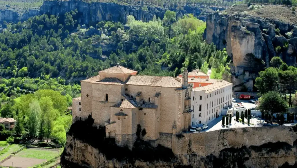 An aerial view of the Parador de Cuenca, a 16th-century former Convent of San Pablo, showcasing its prominent stone church and monastery buildings perched precariously on the edge of a sheer limestone cliff overlooking the Huécar Gorge.