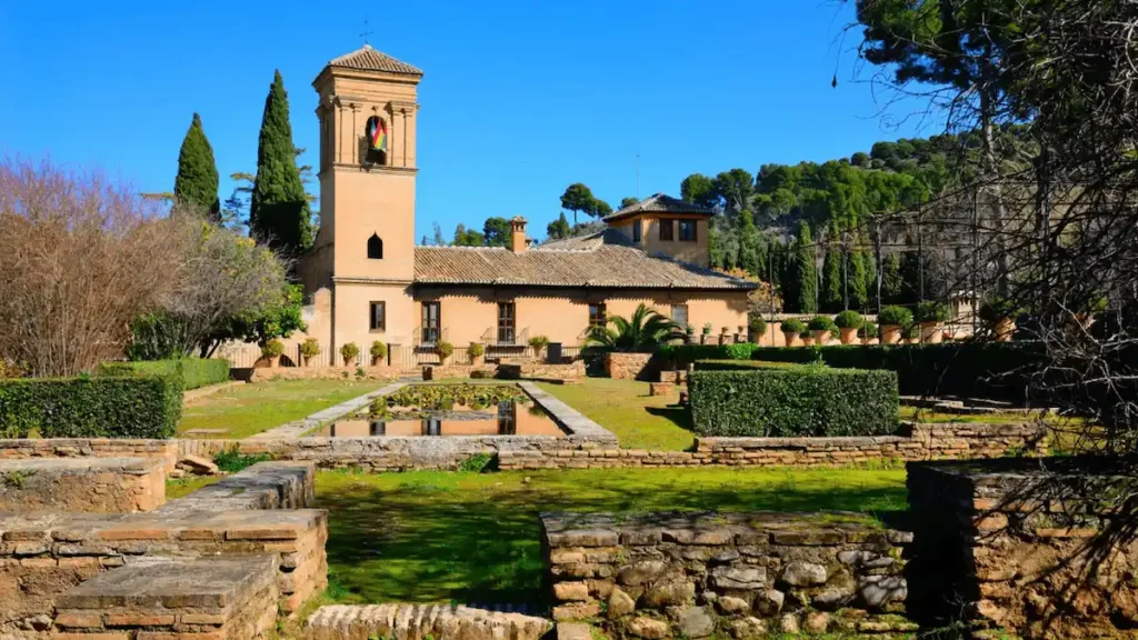 A view of the Parador de Granada, a 15th-century Franciscan convent built upon the remains of a Nasrid palace, featuring its iconic bell tower and a reflecting pool set within the historic gardens of the Alhambra.