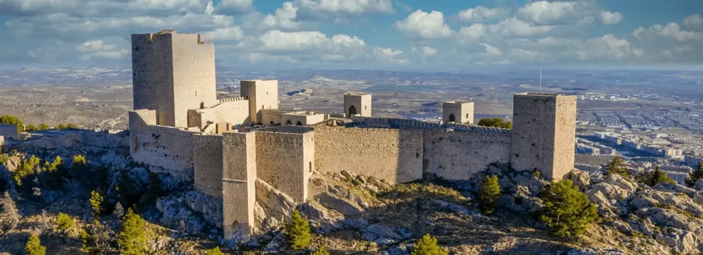 An aerial panorama of the Parador de Jaén, a monumental hotel integrated into the 13th-century Santa Catalina Castle; the image showcases the massive stone defensive walls, a prominent square keep, and several smaller watchtowers perched atop the rugged crest of Santa Catalina Hill.