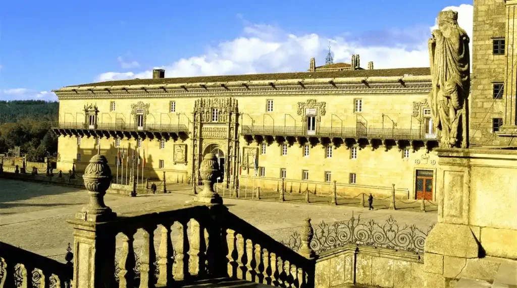 A view from the stone stairs of the Praza do Obradoiro toward the grand 15th-century Plateresque facade of Parador de Santiago - Hostal dos Reis Católicos, showcasing its ornate arched portal and intricate stone carvings originally commissioned by King Ferdinand and Queen Isabella as a royal hospital for pilgrims.