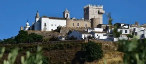 A view of the Pousada Castelo de Estremoz (also known as the Palace of Rainha Santa Isabel) situated atop a hill in the Alentejo region, featuring the 13th-century marble "Tower of Three Crowns" and fortified walls rising above a cluster of traditional white-washed houses.