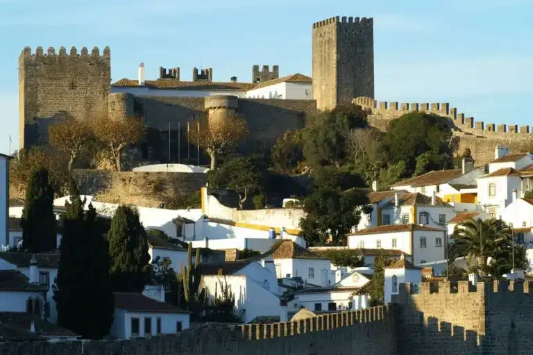 A view of the Pousada Castelo de Óbidos, a premier example of Manueline architecture integrated into a medieval limestone fortress, featuring its massive crenellated keep and defensive curtain walls rising above the traditional white-washed houses of the historic walled village.