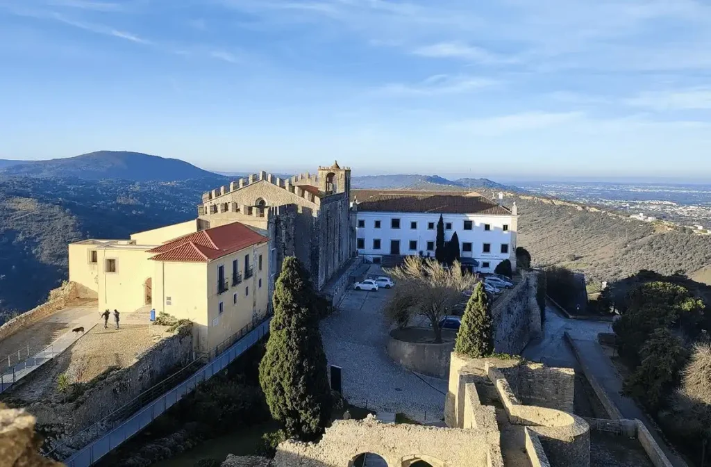 An elevated view of the Pousada Castelo de Palmela, showing the 15th-century Monastery of the Order of Santiago integrated into the medieval hilltop fortress, overlooking the rolling Arrábida hills and the Atlantic coastline.