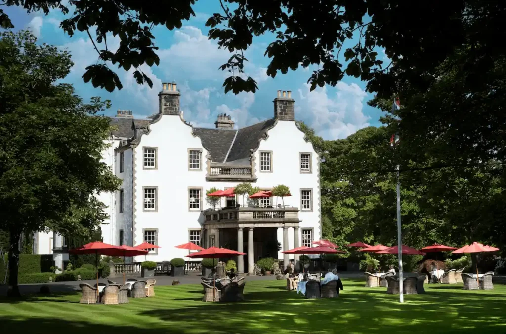 The historic white-harled facade of Prestonfield House in Edinburgh, featuring its signature 17th-century curvilinear gables and red outdoor umbrellas on the lush green lawns of its private 20-acre estate.
