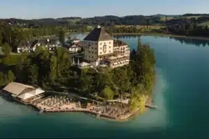 An elevated view of the historic Rosewood Schloss Fuschl, a 15th-century hunting lodge originally built for the Prince-Archbishops of Salzburg, featuring its iconic eggshell-yellow tower and red-roofed wings on a private peninsula overlooking the turquoise waters of Lake Fuschl.