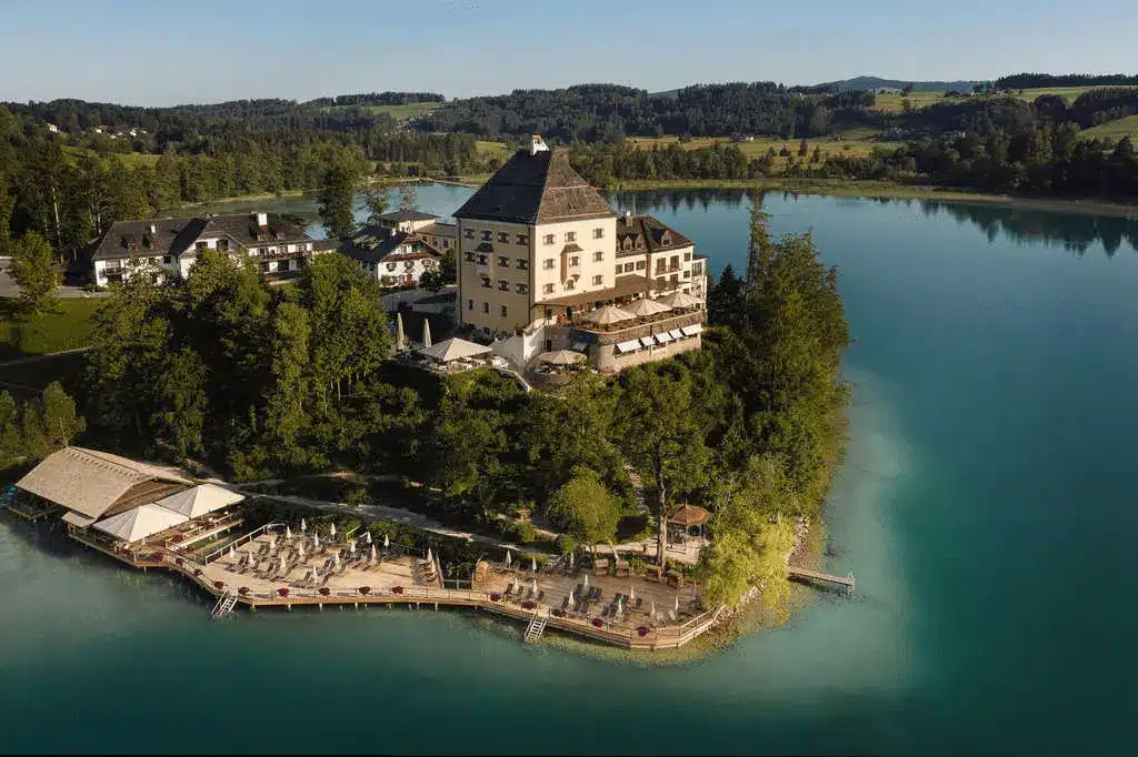 An elevated view of the historic Rosewood Schloss Fuschl, a 15th-century hunting lodge originally built for the Prince-Archbishops of Salzburg, featuring its iconic eggshell-yellow tower and red-roofed wings on a private peninsula overlooking the turquoise waters of Lake Fuschl.