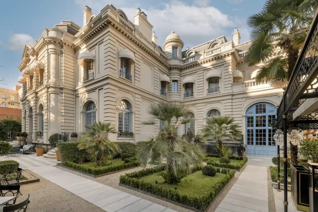 The elegant 19th-century French-style exterior of Santo Mauro Madrid, formerly the private residence of the Duke of Santo Mauro, featuring neoclassical stone architecture and a manicured garden with palm trees and boxwood hedges.