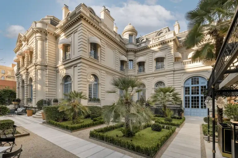 The elegant 19th-century French-style exterior of Santo Mauro Madrid, formerly the private residence of the Duke of Santo Mauro, featuring neoclassical stone architecture and a manicured garden with palm trees and boxwood hedges.