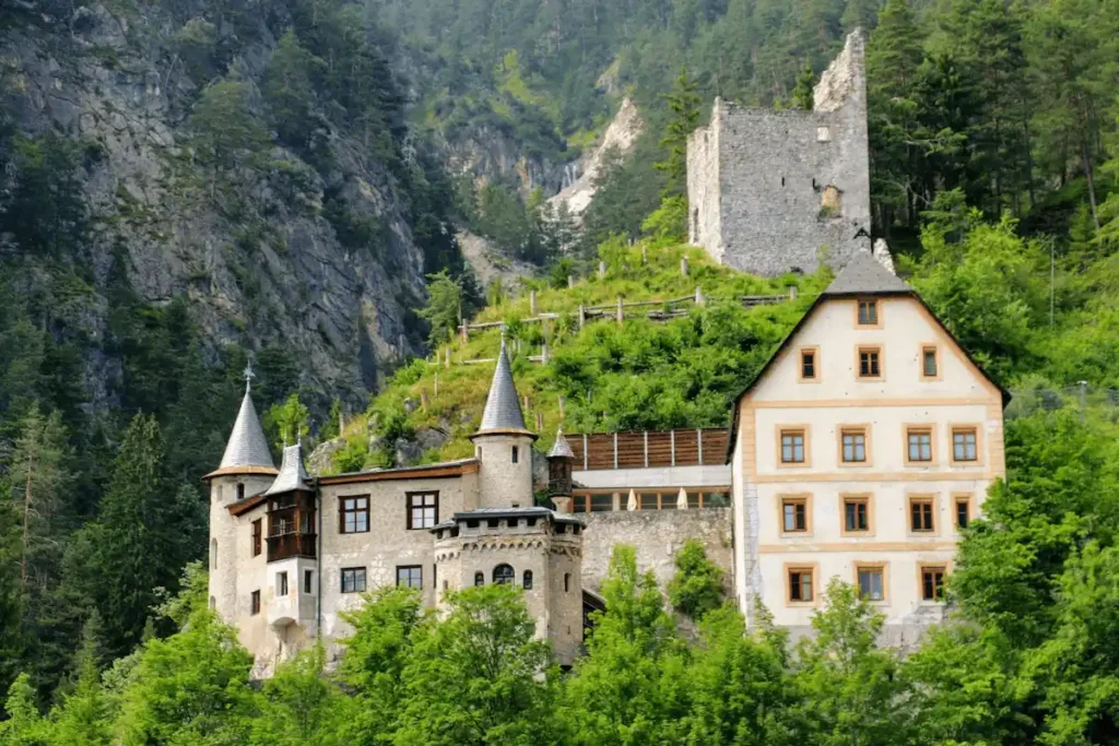 A view of the historic Schloss Hotel Fernsteinsee in the Austrian Alps, featuring its fairy-tale turrets and a yellow-walled manor house set against a sheer mountain cliff, with the stone ruins of the 13th-century Sigmundsburg Castle visible on the forested hilltop above.