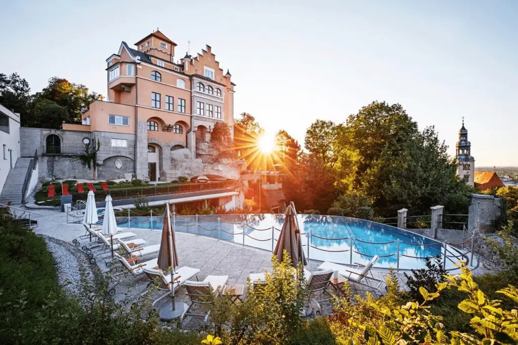 A sunset view of Hotel Schloss Mönchstein, a 14th-century fairy-tale castle in Salzburg, featuring its iconic stepped gables and peach-colored facade rising above a modern glass-edged infinity pool with panoramic views over the city.