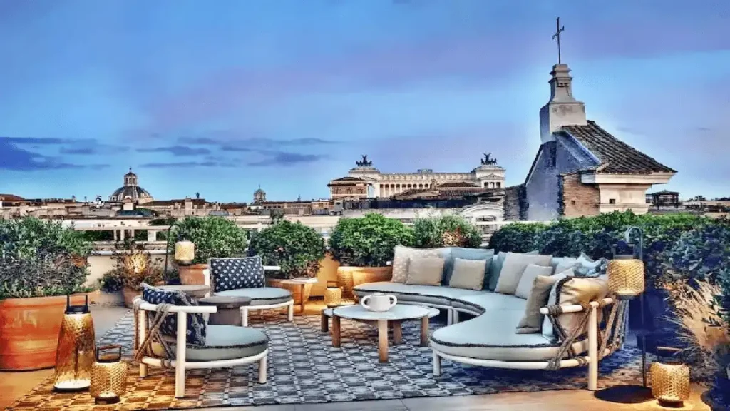 The sustainable luxury rooftop terrace of Six Senses Rome, featuring contemporary outdoor lounge seating and panoramic views of the Roman skyline, including the neighboring San Marcello al Corso Church and the Altare della Patria.