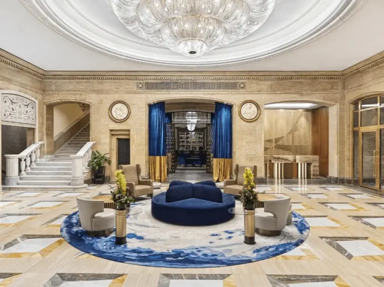 The opulent lobby of the Sofia Balkan Palace, featuring a grand crystal chandelier, marble flooring with geometric patterns, and elegant blue velvet seating arrangements.